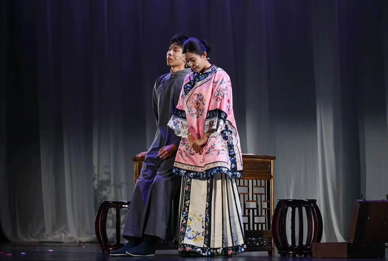 一位穿灰色长衫的演员坐在木桌旁，身旁穿粉色绣花服装的演员低头靠近他
