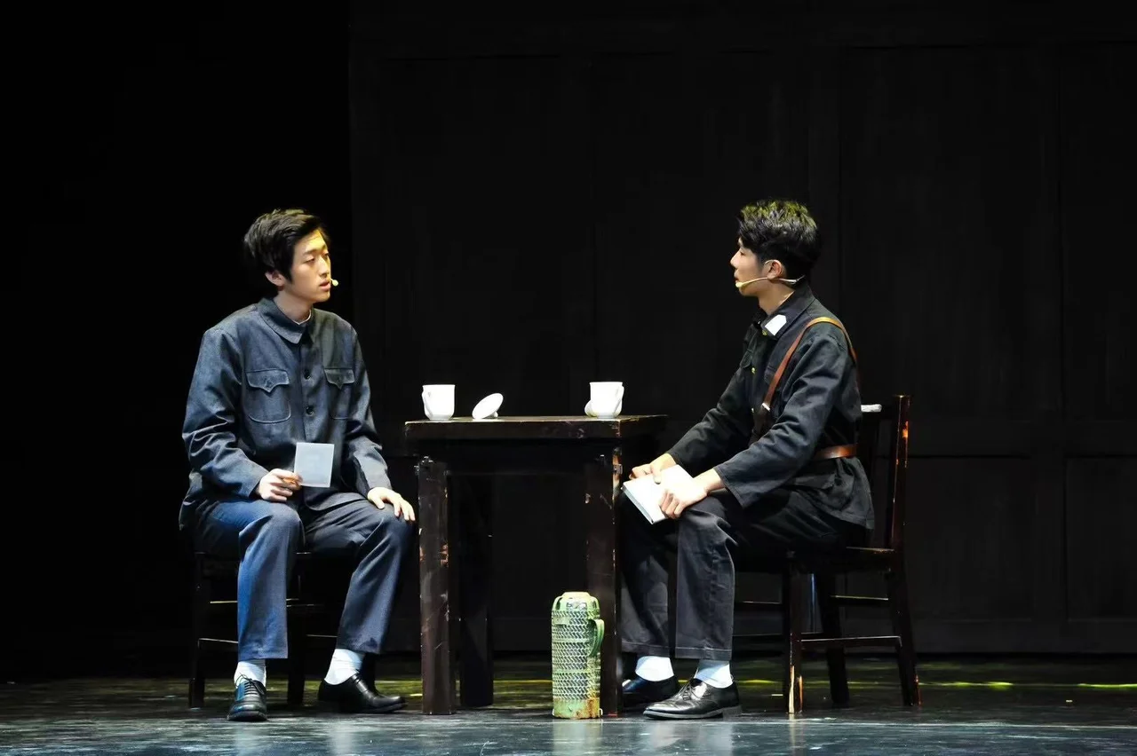两位穿制服的演员隔桌相对而坐，手持纸张，桌上放着白色杯子