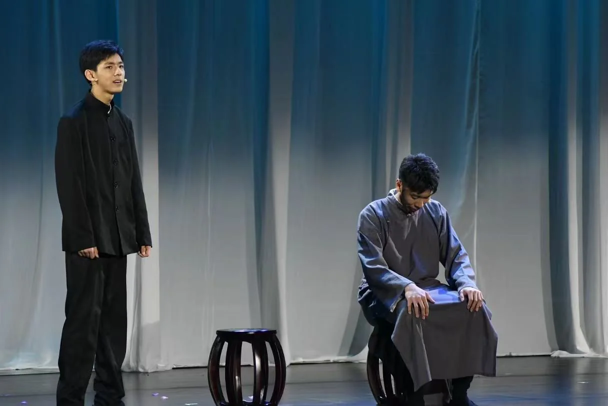 穿黑色服装的演员站在左侧，穿灰色长衫的演员低头坐在右侧圆凳上