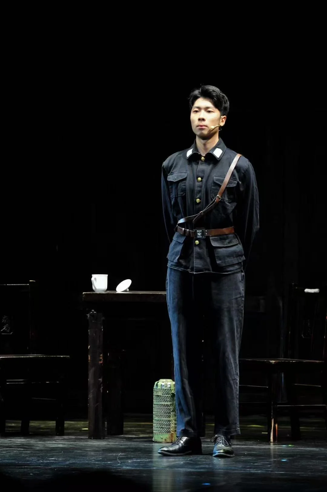 穿深色制服、系腰带和斜肩带的演员站在舞台上，身旁桌上放着白色杯子