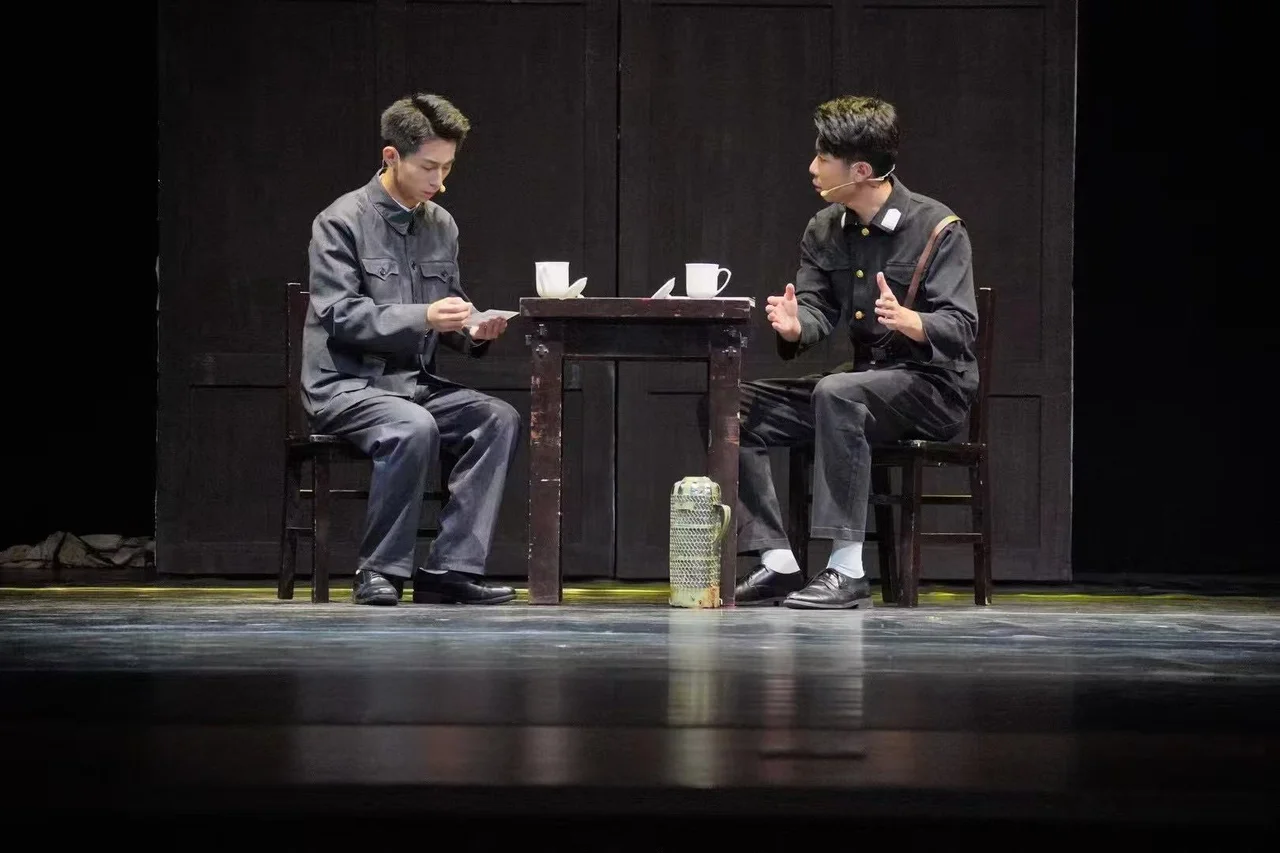 两位穿制服的演员坐在桌子两侧，左侧演员看着纸张，右侧演员张开双手说话