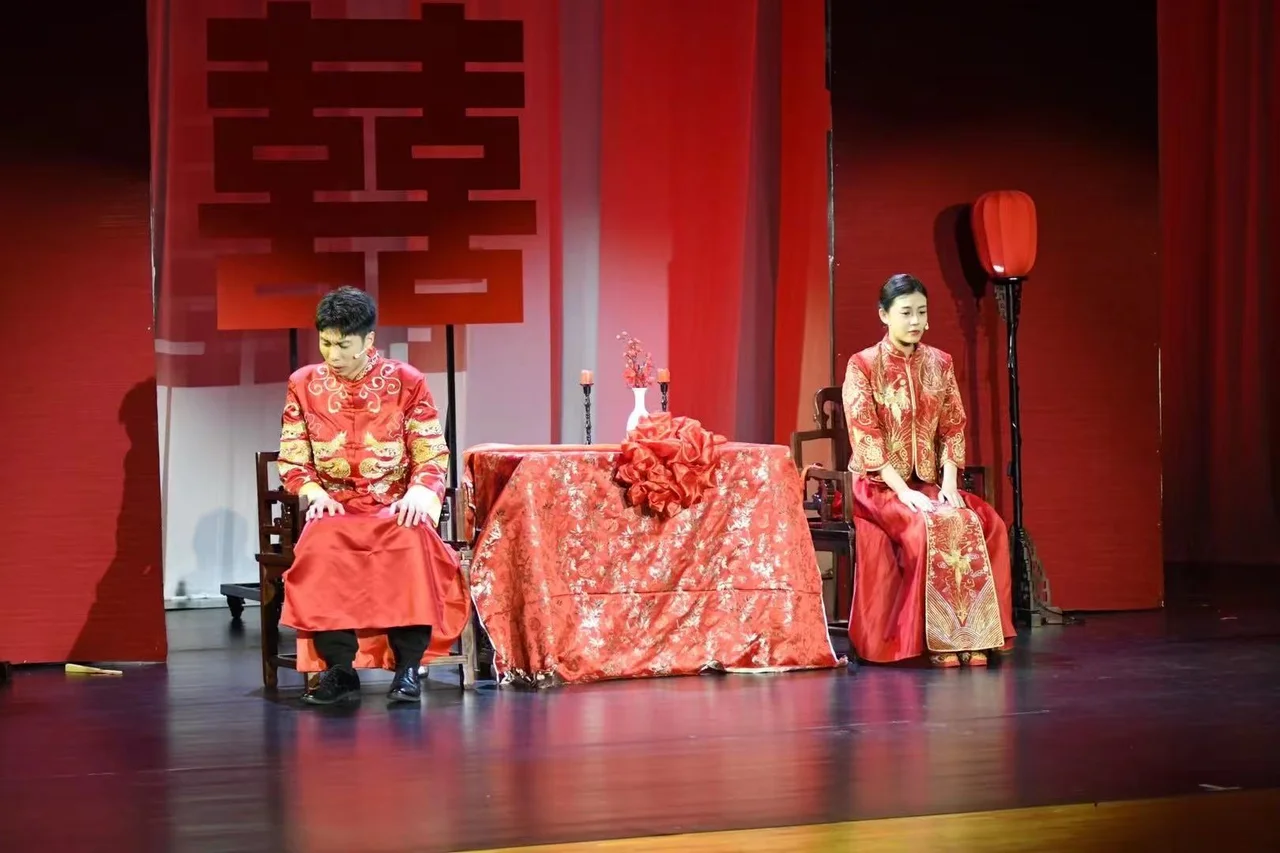 两位身穿红色婚服的演员坐在桌子两侧，舞台背景是红色囍字和灯笼