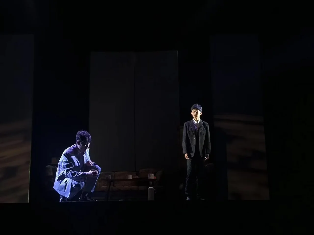 暗色舞台上，一位演员蹲在左侧，另一位穿深色西装的演员站在右侧灯光中