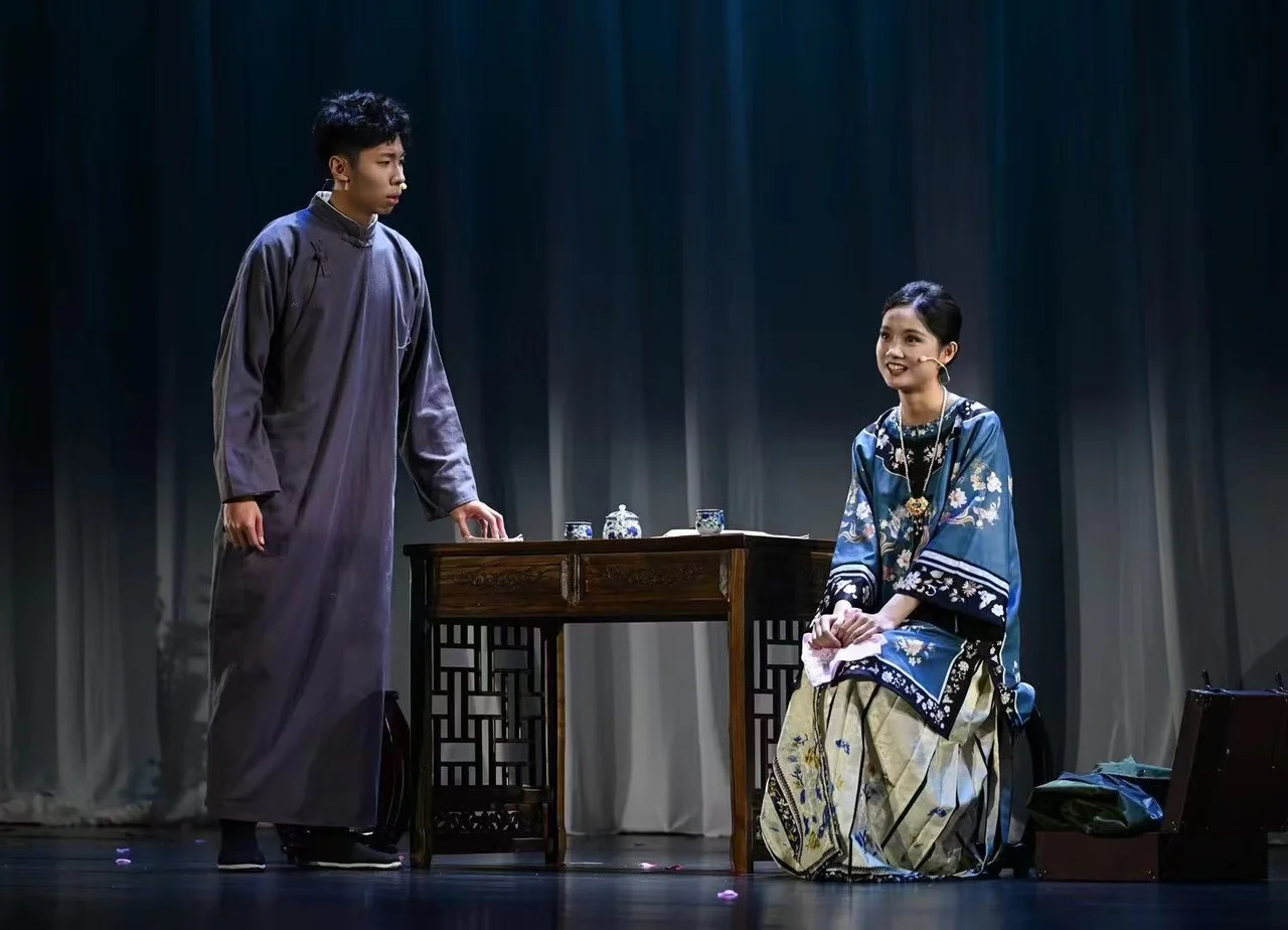 穿灰色长衫的演员站在木桌左侧，穿蓝色绣花服装的演员坐在右侧，桌上摆着茶具