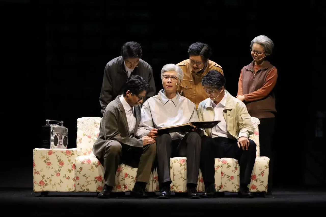《与光同行》舞台上,灰发演员坐在花纹沙发中央翻看相册,其他演员围在身旁
