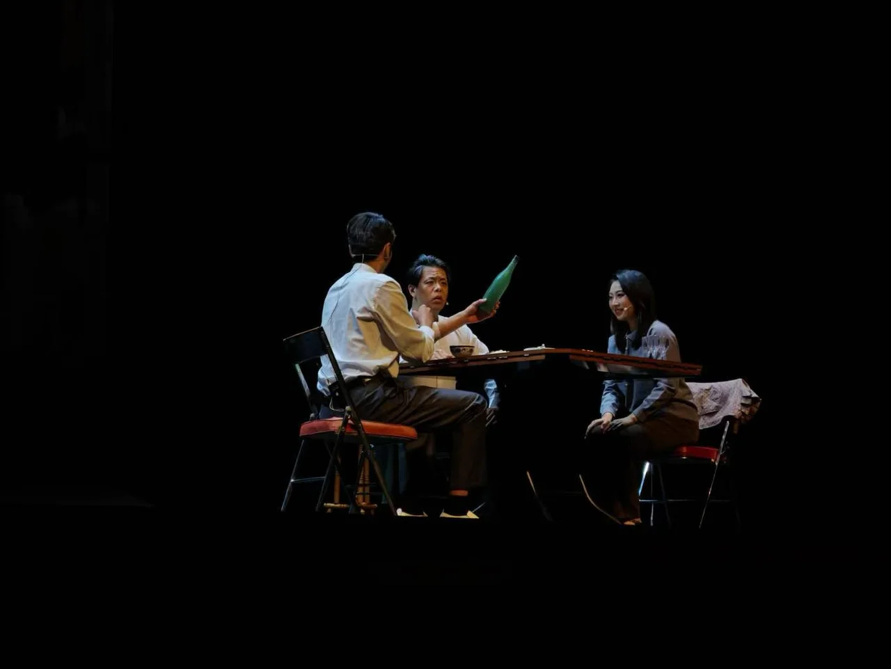 《与光同行》舞台上,三位演员围坐桌旁,其中一人举起绿色瓶子